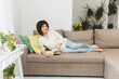 © inna717 - Young Asian woman sitting on a modern sofa, relaxing in her living room, reading a book at home