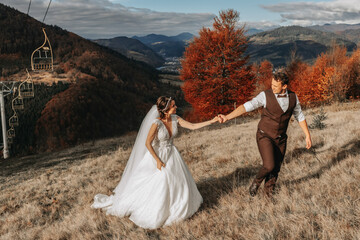 Wedding couple walks in the mountains. The groom leads the bride by the hand. The dress of the bride develops in the wind.