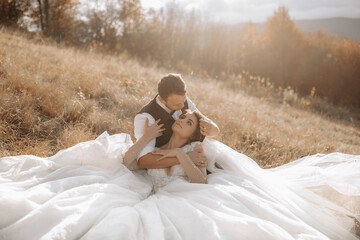 Wall Mural - portrait of a stylish groom with a bride on a background of autumn dry grass. the concept of a rural wedding in the mountains, happy bohemian newlyweds. the bride and groom are lying on the grass