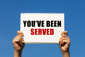 You've been served text on notebook paper held by 2 hands with isolated blue sky background. This message can be used as business concept about you've been served.