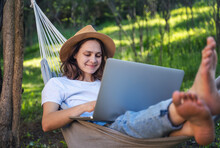 Young Cheerful Woman Working Online Using Laptop Lying In Hammock In Summer Garden