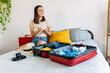 © Xavier Lorenzo - Young beautiful woman sitting on bed packing her suitcase for travel vacation. Smiling female traveler writing planner summer trip on notebook. Summer vacation lifestyle concept.