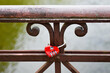 A red lock in the form of a heart hanging on a brown decorative forged element of the bridge railing against on the background of river water