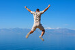 Young man jumping on rock with his hands up blue ocean mountain peaks horizon. Active holiday adventure, tourism action, healthy summer joy, Fun activity lifestyle. Crazy adult guy fly from climb.