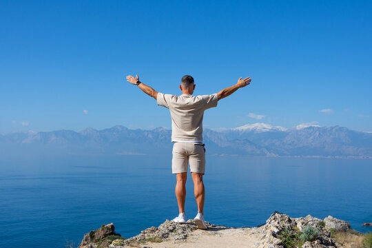 Man stands with arms raised on rock blue ocean mountain peaks horizon. Active holiday adventure, tourism action, healthy summer joy, Fun activity lifestyle. Crazy adult guy fly from climb.