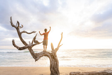 Canvas Print - Horizontal of a man enjoying a beautiful sunset standing with his arms raised on a dry tree trunk in the middle of the beach. 