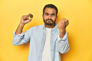 Wall Mural - Young Latino man holding a Bitcoin coin, yellow studio background, showing fist to camera, aggressive facial expression.