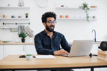 Wall Mural - Joyful bearded man in glasses operating modern laptop while sitting at writing desk in spacious dining room. Smiling indian freelancer using flexible schedule while spending working hours at home.