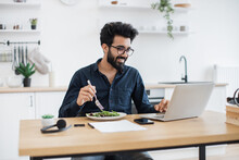 Smiling Mature Man In Business Casual Clothes Eating Healthy Salad While Looking At Laptop Screen In Kitchen Interior. Indian Remote Employee Having Lunch Break While Doing Home-based Job At Midday.