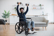 Joyful arabian person with physical disability holding dumbbells in raised arms during strength training at home. Active wheelchair user building muscles while experimenting with workout routine.
