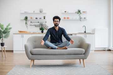 Full length view of mature person doing yoga exercise while sitting with legs crossed on couch in dining room. Barefoot indian man in spectacles relaxing with closed eyes after working day at home.