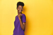 © Asier - African woman in violet tee, yellow studio scene, points with thumb finger away, laughing and carefree.