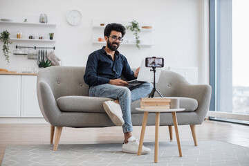 Arabian male blogger in glasses and casual clothes recording training video on modern smartphone in cozy studio. Handsome positive man holding portable laptop on legs while sitting on grey sofa.