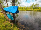 Fototapeta  - Two blue kayaks are situated within a tree, awaiting their next adventure. The tree hangs over an idyllic pond setting with green leafy trees lining the shore. Cloudy blue skies are overhead.