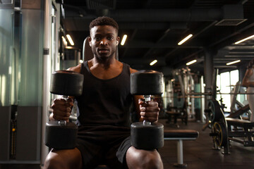 athletic african american man trains in dark gym, athletic guy lifts heavy dumbbells in fitness club