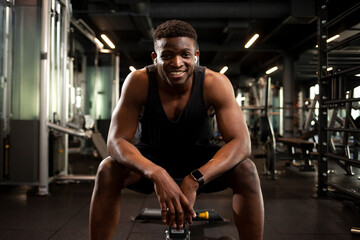 Wall Mural - young athletic african american man sits in dark gym and smiles, athletic guy rests in fitness club