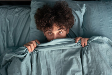 Handsome surprised guy covering half of face with blanket, scared man peeking from duvet, afraid of night monsters, top view