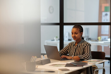 Aufkleber - Young serious African American female designer typing on laptop keyboard while sitting by workplace in office and working over new project