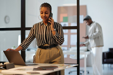 Poster - Young confident businesswoman or designer with smartphone by ear standing by workplace with laptop and communicating with client