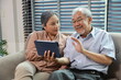 © feeling lucky - Happy smiling asian senior couple sitting on sofa and using tablet while online video call with friend or relative cousin at home living room. Internet information technology and lifestyle concept.