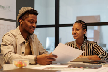 Aufkleber - Young smiling businessman with papers showing and explaining data to female colleague sitting by desk next to him during working meeting