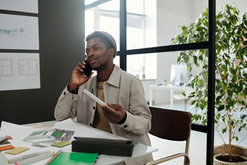 Aufkleber - Young confident black man in casualwear talking to colleague on mobile phone while sitting by desk with sketches, catalog and office supplies