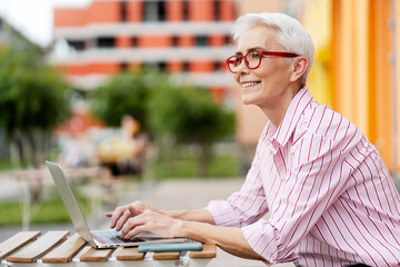 Wall Mural - Portrait of smiling positive senior woman wearing modern red glasses, working at computer, using laptop outdoors, looking away, copy space. Successful freelancer, workplace. Remote job concept