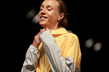 Wall Mural - Young fitness girl getting ready for a workout at the stadium. Attractive slender blonde in a yellow-gray sports jacket is focused on training. Active lifestyle. Close up shot.