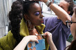 The makeup artist creates a stage makeup for an African woman in a green fur coat, a stylish hairdo, a brush applied highlighter to the cleavage area, the hairdresser completes the hair style