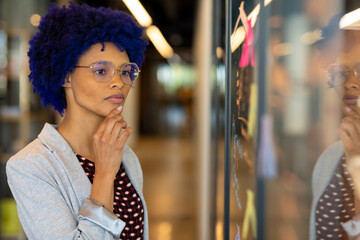 Thoughtful biracial casual businesswoman with blue afro brainstorming at glass wall in office