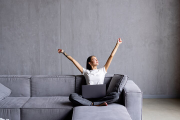 Beautiful young woman is using a laptop and smiling with win gesture while sitting on the sofa at home