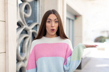 Poster - young pretty woman looking surprised and shocked, with jaw dropped holding an object with an open hand on the side