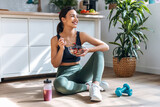 Fototapeta Lawenda - Athletic woman eating a healthy bowl of muesli with fruit sitting on floor in the kitchen at home