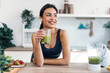 Athletic smiling woman drinking smothie while listening music with earphones in the kitchen at home.