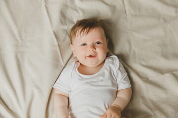 charming blue-eyed 6-month-old baby lies in bed in a white bodysuit. view from above