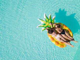 Wall Mural - Top view of two girls relaxing on inflatable big pineapple in the pool. Aerial view of ladies relaxing on the floating mattress.