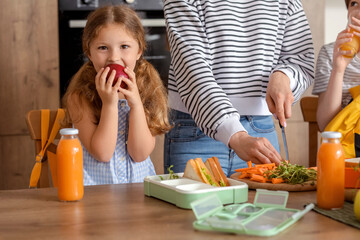 Wall Mural - Little girl eating apple while her mother preparing school lunch in kitchen