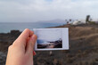 A young woman's hand holds a polaroid photo in front of the view where it was taken