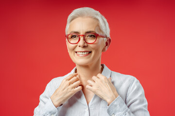 Wall Mural - Smiling gray haired senior woman wearing stylish eye wear isolated on red