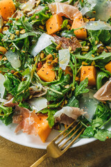 Sticker - top down view of arugula salad with cheese, cantaloupe, prosciutto, pistachios on white plate. golden fork, gold background