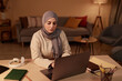 © Seventyfour - Young serious Muslim businesswoman sitting by workplace in living room and looking at laptop screen while scrolling through online data