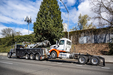 Powerful black big rig tow semi truck towing broken big rig semi truck without semi trailer driving on the highway road at sunny day