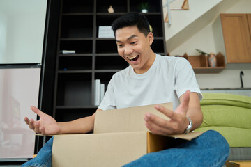 Young asian man unpacking  awaited parce looking inside, sitting at sofa, satisfied happy customer opening cardboard box with online store order, good shipping delivery service