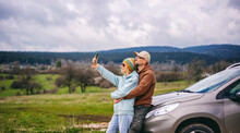 Romantic Middle Age Couple Hugging Standing Next To Cars During Auto Travel In Countryside. Taking A Selfie Using A Smartphone