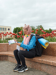 An attractive young tourist in a blue shirt is exploring a new city. A girl with long pink hair holds a paper map of the city scheme in her hands. Travel around Europe.