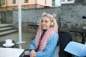 An attractive young tourist in a blue shirt drinks coffee and listens to music with headphones. A girl with long pink hair is having lunch in a cafe. Travel around Europe.