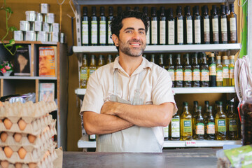 Latin man greengrocer in an organic grocery shop behind the counter with his arms crossed looking at camera