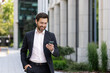 © Tetiana - Mature adult male boss walking outside office building, successful businessman typing message and browsing online pages happily, investor in business suit with smartphone