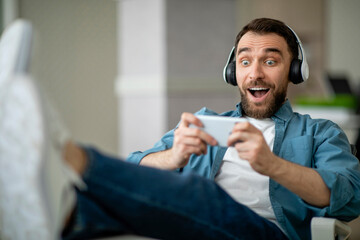 Aufkleber - Cheerful Millennial Guy In Wireless Headphones Playing Video Games On Smartphone