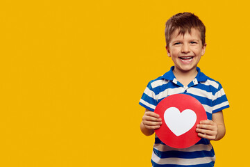 Happy cute boy in blue striped polo, showing heart icon for social networks while smiling against yellow background with empty space